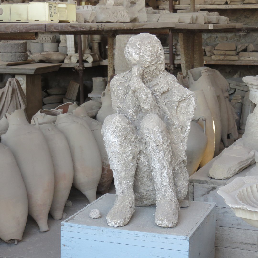 Plaster cast of a seated Pompeii victim on display among amphorae in a storeroom